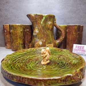 May include: A set of ceramic tree stump-shaped serving pieces, including a pitcher, four cups, and a large platter. The pieces are decorated with a green and brown glaze, and the platter features a small squirrel figurine in the center.