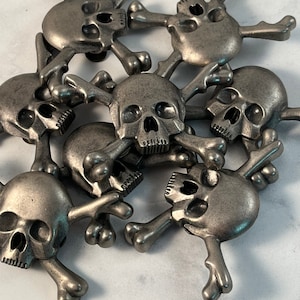 Pode incluir: Um grupo de dez pingentes de prata com formato de caveira e ossos cruzados. Os pingentes são de metal e têm um acabamento desgastado. Todos estão em uma pilha, com alguns sobrepostos.