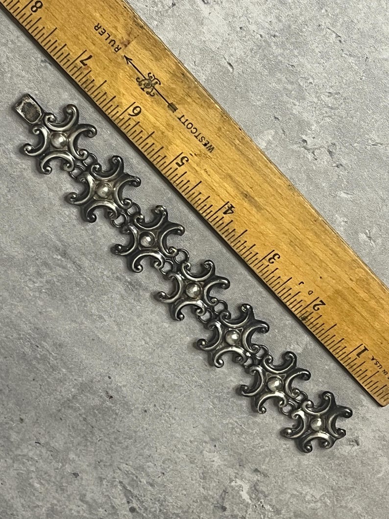 Peut inclure: Un bracelet en argent avec un motif r&eacute;p&eacute;t&eacute; de croissants de lune stylis&eacute;s et de petites perles rondes. Le bracelet mesure environ 15 cm de long et est mesur&eacute; contre une r&egrave;gle en bois.