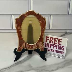 May include: A small, oval-shaped mirror with a brown and gold frame. The frame has a swirl pattern and the mirror is on a black stand.