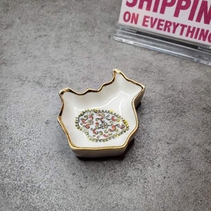 May include: A small white ceramic dish shaped like a fish with a gold rim. The dish has a colorful floral design in the center.