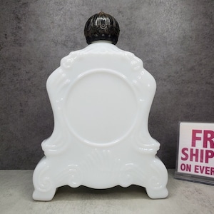 May include: White porcelain bottle with a decorative, ornate design. The bottle has a round, white base and a black metal top.