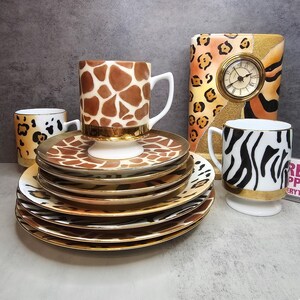 Puede incluir: Un juego de vajilla de cerámica con diseños de animales. El juego incluye platos, tazas y un reloj decorativo. Las tazas presentan diseños de jirafa, leopardo y cebra, con detalles dorados. Los platos tienen estampados de jirafa y leopardo.