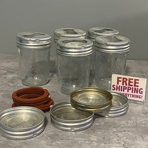 Op de afbeelding: Zes glazen potten met zilveren deksels en rubberen ringen. De potten zijn gerangschikt in twee rijen van drie. Er zijn ook drie zilveren deksels en een stapel rubberen ringen op een grijs oppervlak. Een bordje met de tekst "GRATIS VERZENDING ALLES!"