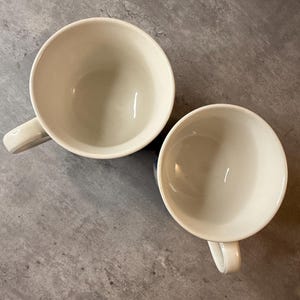 May include: Two off-white ceramic coffee mugs with handles, set against a gray background. The mugs are empty and appear to be clean. The mugs are round and have a simple, classic design.