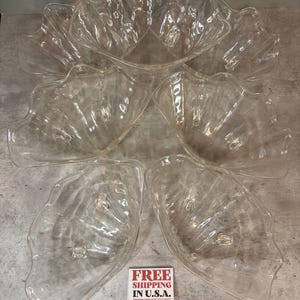 Puede incluir: Un conjunto de cinco cuencos de cristal transparente con forma de concha marina, con bordes festoneados y pequeños pies de cristal en la parte inferior. Los cuencos están dispuestos en grupo, con uno grande arriba y cuatro más pequeños debajo.