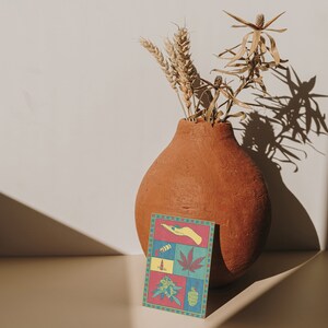 May include: A terracotta vase holds dried wheat and plants, casting a shadow on a light-colored surface. A colorful card with a cannabis theme is in front of the vase. The card features various cannabis-related illustrations in a grid pattern.
