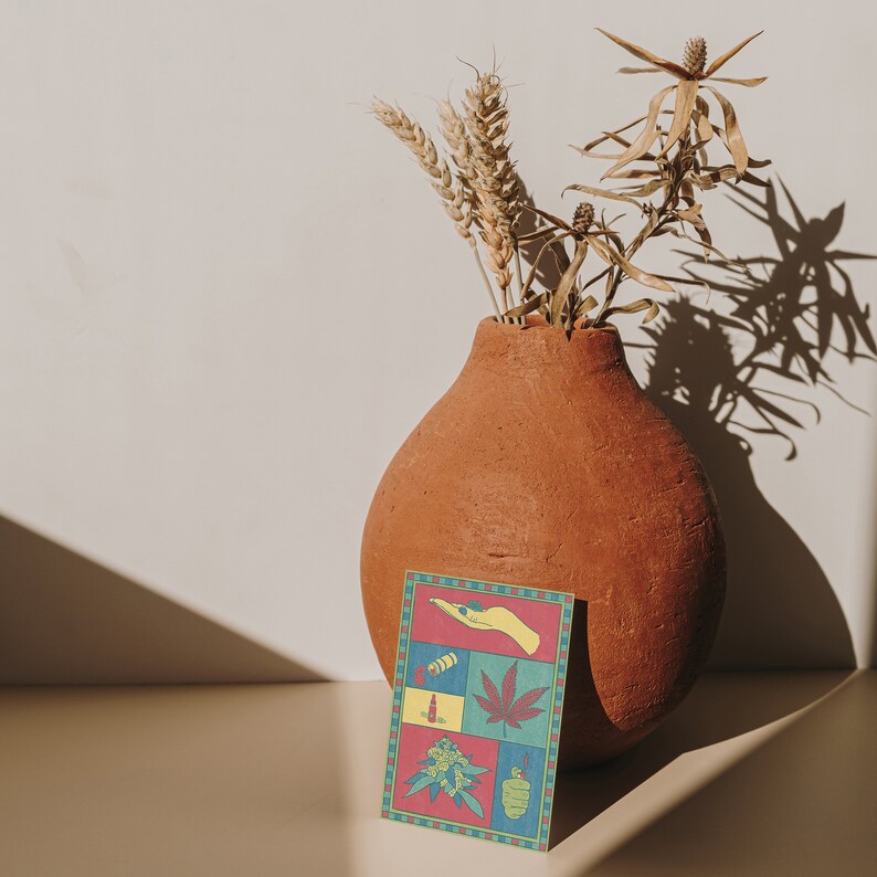 May include: A terracotta vase holds dried wheat and plants, casting a shadow on a light-colored surface. A colorful card with a cannabis theme is in front of the vase. The card features various cannabis-related illustrations in a grid pattern.