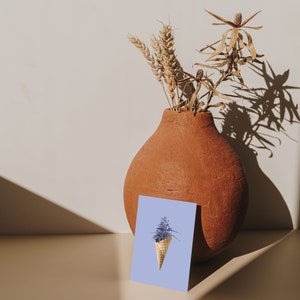 May include: A light blue card with a purple flower in a waffle cone on a light blue background. The card is sitting in front of a terracotta vase with dried flowers.