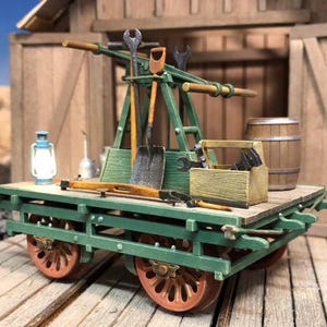 May include: A green wooden model of a railway maintenance cart with a variety of tools, including shovels, wrenches, and a lantern. The cart has two wheels on each side and a wooden barrel on the back.