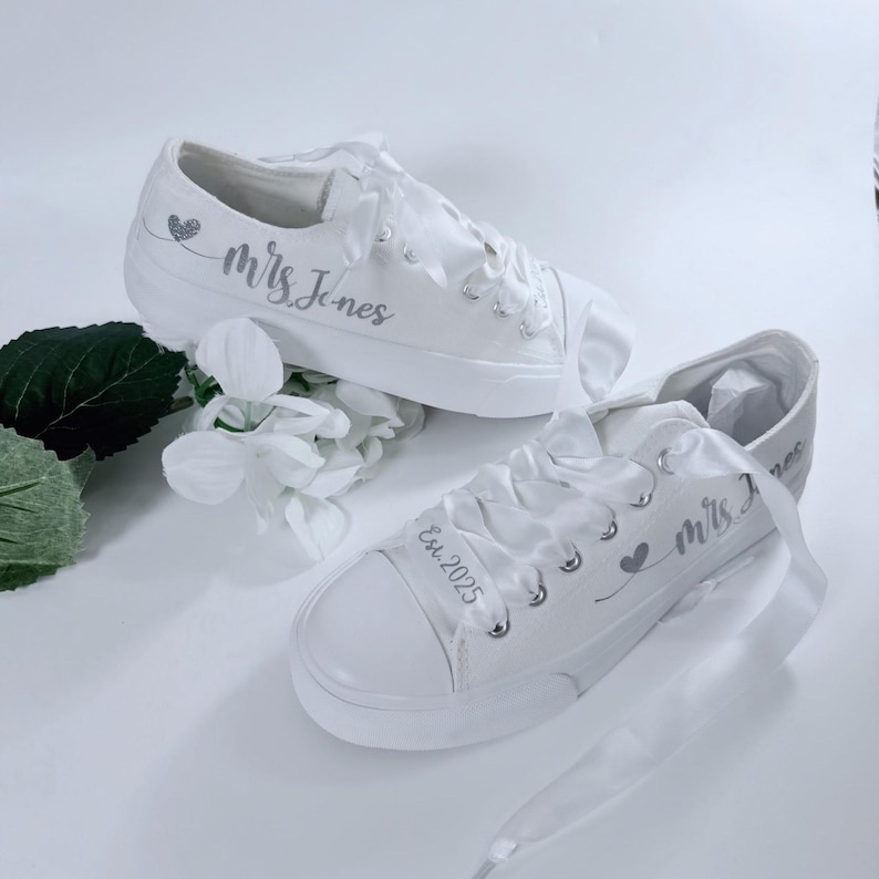 Flat Slip On Bride, two white canvas sneakers with satin laces. One shoe has the text 'Mrs. last name marriage' and a small heart design. The other shoe has the text 'Est. time' and a small heart design.