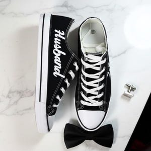 K&ouml;nnte beinhalten: Schwarze Leinenschuhe mit wei&szlig;en Schn&uuml;rsenkeln und Sohlen. Auf einem Schuh steht das Wort "Husband" in wei&szlig;er Schrift. Eine schwarze Fliege und silberne Manschettenkn&ouml;pfe sind ebenfalls vorhanden.