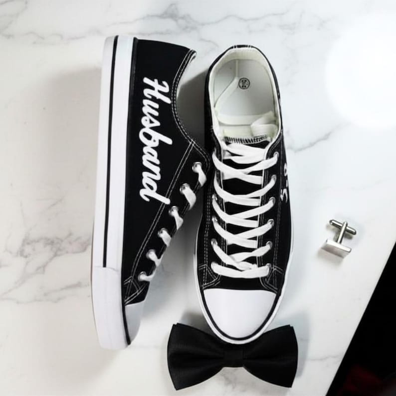 K&ouml;nnte beinhalten: Schwarze Leinenschuhe mit wei&szlig;en Schn&uuml;rsenkeln und Sohlen. Auf einem Schuh steht das Wort "Husband" in wei&szlig;er Schrift. Eine schwarze Fliege und silberne Manschettenkn&ouml;pfe sind ebenfalls vorhanden.