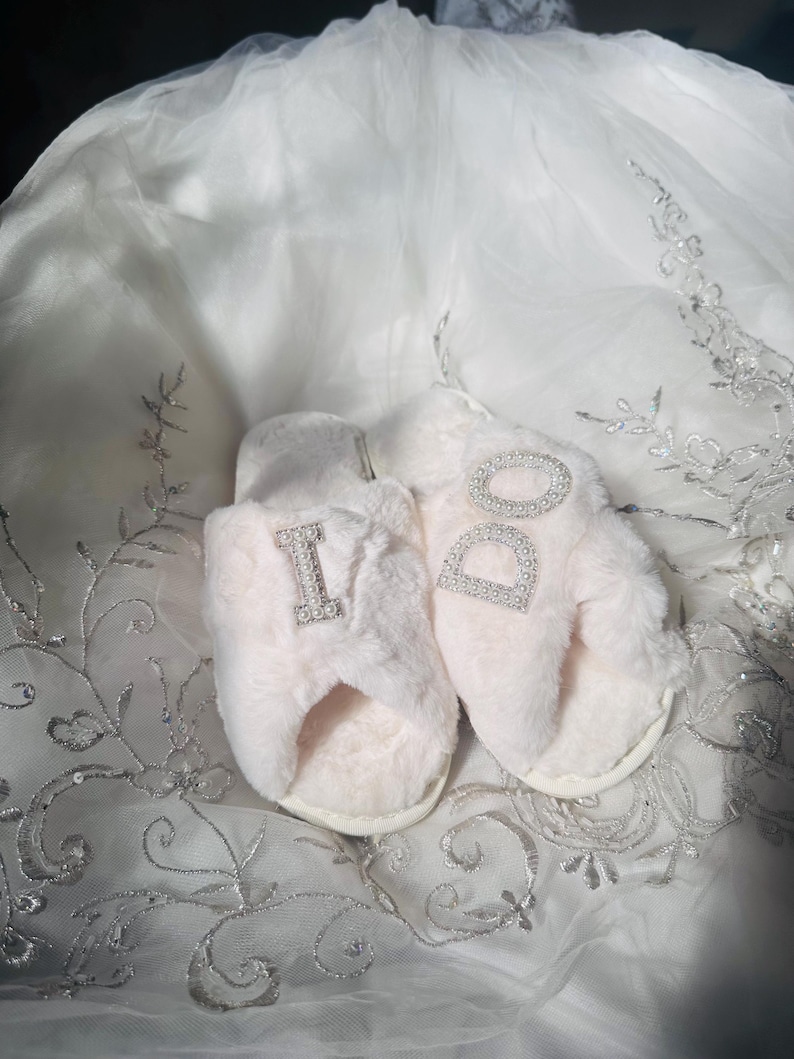 Pode incluir: Um par de chinelos rosa claro e fofos com as palavras "I DO" em strass brilhantes. Os chinelos est&atilde;o sobre um tecido branco bordado, sugerindo um tema nupcial.