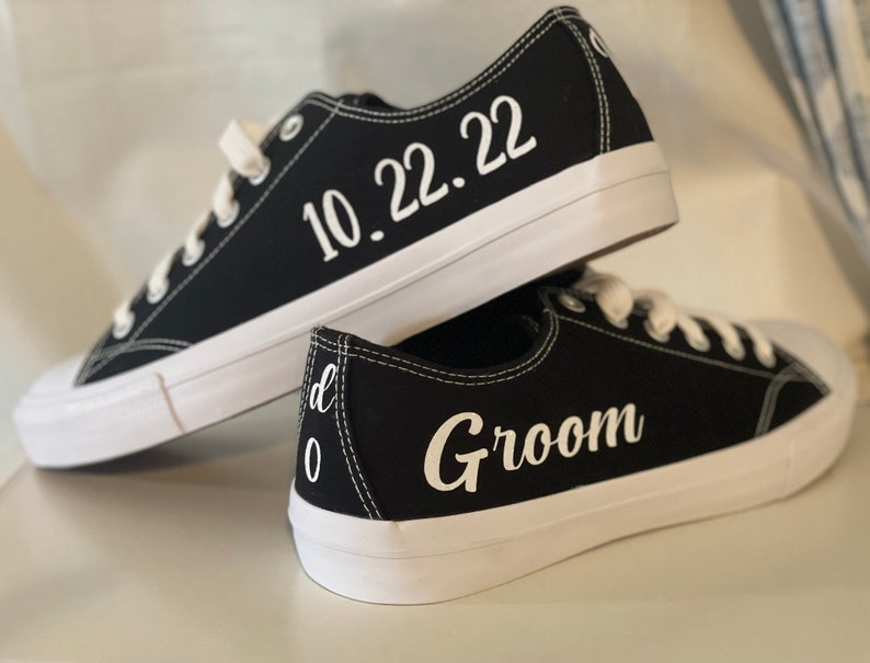 K&ouml;nnte beinhalten: Schwarze Leinenschuhe mit wei&szlig;en Sohlen und Schn&uuml;rsenkeln. Ein Schuh hat das Datum "10.22.22" in Wei&szlig; aufgedruckt. Der andere Schuh tr&auml;gt den Schriftzug "Groom" in wei&szlig;er Schrift.