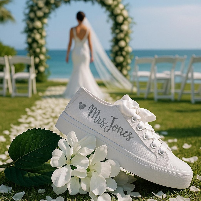 Pode incluir: T&ecirc;nis de lona brancos com cadar&ccedil;os de fita de cetim, com o texto "Mrs Jones" em prata e um pequeno cora&ccedil;&atilde;o. Os sapatos est&atilde;o colocados sobre um leito de flores brancas e grama, com uma noiva e um arco de casamento ao fundo.
