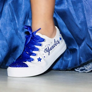 Puede incluir: Zapatillas blancas de lona con cordones de cinta azul y pedrer&iacute;a azul. Las zapatillas est&aacute;n decoradas con estrellas plateadas de purpurina y el nombre "Yanelis" est&aacute; escrito en cursiva azul.