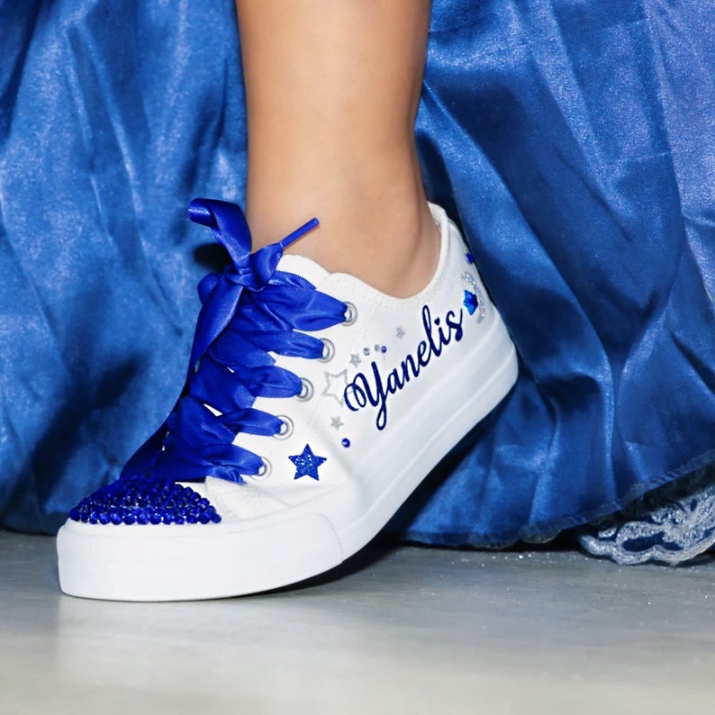 Puede incluir: Zapatillas blancas de lona con cordones de cinta azul y pedrer&iacute;a azul. Las zapatillas est&aacute;n decoradas con estrellas plateadas de purpurina y el nombre "Yanelis" est&aacute; escrito en cursiva azul.