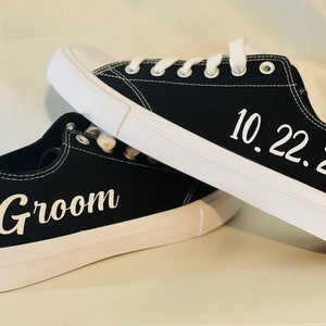 K&ouml;nnte beinhalten: Schwarze Leinenschuhe mit wei&szlig;en Sohlen und Schn&uuml;rsenkeln. Auf einem Schuh steht das Wort "Groom" in wei&szlig;er Schrift, auf dem anderen das Datum "10.22.22". Ein stilvolles Hochzeitsgeschenk.