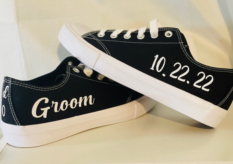 K&ouml;nnte beinhalten: Schwarze Leinenschuhe mit wei&szlig;en Sohlen und Schn&uuml;rsenkeln. Auf einem Schuh steht das Wort "Groom" in wei&szlig;er Schrift, auf dem anderen das Datum "10.22.22". Ein stilvolles Hochzeitsgeschenk.