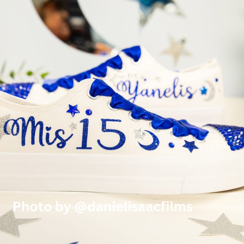 Puede incluir: Zapatillas blancas de lona con detalles de purpurina azul y una cinta azul. Las zapatillas tienen un dise&ntilde;o de media luna y estrellas y el texto "Mis 15" en una zapatilla y "Yanelis" en la otra.