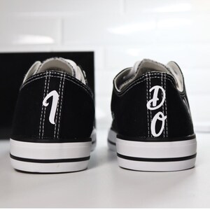 K&ouml;nnte beinhalten: Paar schwarze Leinenschuhe mit wei&szlig;en Sohlen und Schn&uuml;rsenkeln. Die Fersen zeigen die wei&szlig;en Buchstaben "1" und "DO". Die Schuhe haben ein klassisches, flaches Design.