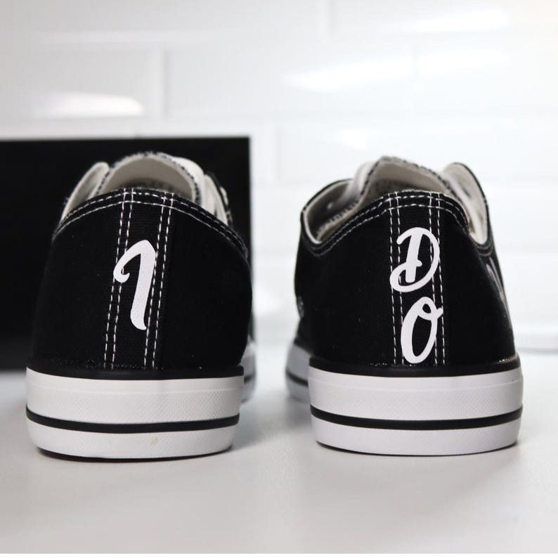 K&ouml;nnte beinhalten: Paar schwarze Leinenschuhe mit wei&szlig;en Sohlen und Schn&uuml;rsenkeln. Die Fersen zeigen die wei&szlig;en Buchstaben "1" und "DO". Die Schuhe haben ein klassisches, flaches Design.