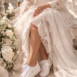 Pode incluir: Uma noiva com um vestido de noiva de renda branca e t&ecirc;nis brancos. Os t&ecirc;nis s&atilde;o personalizados com o texto "Mrs Jones". O vestido apresenta mangas compridas e decote em V, com detalhes florais e acabamento em renda.