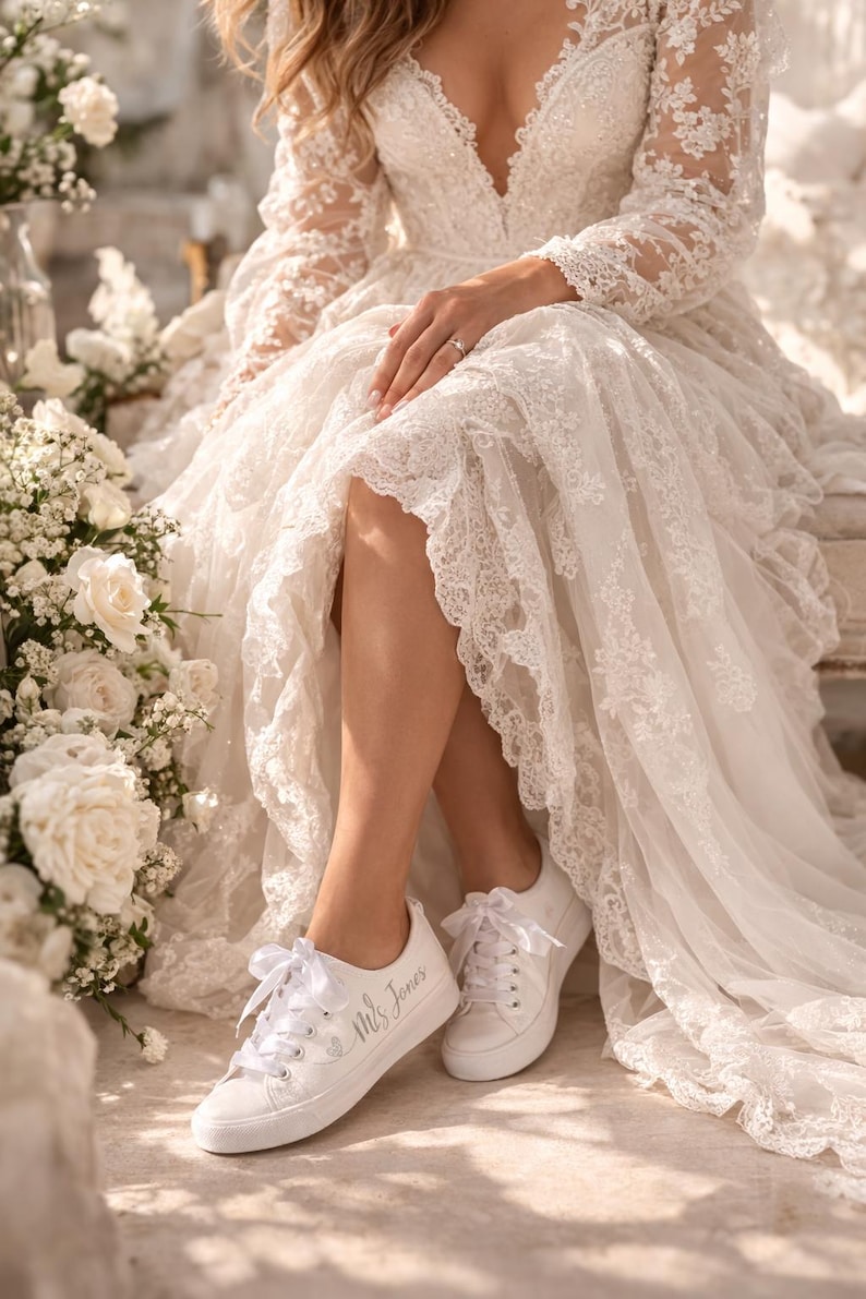 Pode incluir: Uma noiva com um vestido de noiva de renda branca e t&ecirc;nis brancos. Os t&ecirc;nis s&atilde;o personalizados com o texto "Mrs Jones". O vestido apresenta mangas compridas e decote em V, com detalhes florais e acabamento em renda.