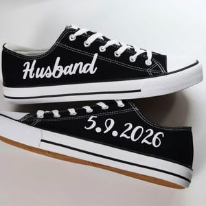K&ouml;nnte beinhalten: Schwarze Leinenschuhe mit wei&szlig;en Schn&uuml;rsenkeln und Sohlen. Auf einem Schuh steht das Wort "Husband" in wei&szlig;er Schrift, auf dem anderen das Datum "5.9.2026". Die Schuhe haben ein klassisches Low-Top-Design.