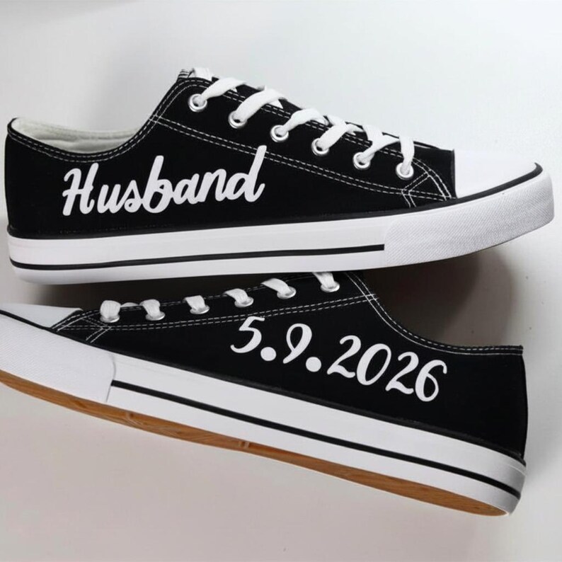 K&ouml;nnte beinhalten: Schwarze Leinenschuhe mit wei&szlig;en Schn&uuml;rsenkeln und Sohlen. Auf einem Schuh steht das Wort "Husband" in wei&szlig;er Schrift, auf dem anderen das Datum "5.9.2026". Die Schuhe haben ein klassisches Low-Top-Design.