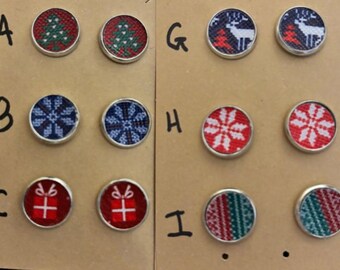 May include: A collection of Christmas-themed round buttons with silver metal rims. Designs include Christmas trees, snowflakes, reindeer, and gift boxes in red, green, and blue patterns. The buttons are arranged on a brown surface.