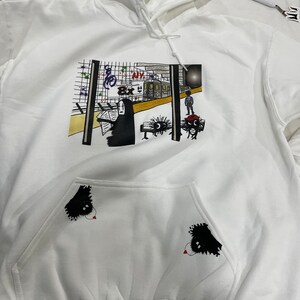 Op de afbeelding: Witte hoodie met een zwart-witte grafische print van een metroscène met de tekst "NY" op een trein. De grafiek bevat ook een persoon met een zwarte cape en twee zwarte figuren met rode ogen. De hoodie heeft een zwarte zak met een zwart-witte grafische print van een figuur met rode ogen.