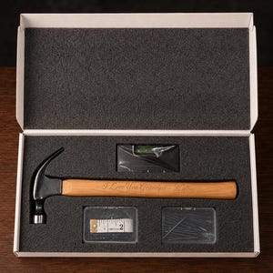 May include: A white gift box with black foam inserts containing a claw hammer with a wooden handle engraved with the text I Love You Grandpa, a small spirit level, a white measuring tape, and a clear plastic container filled with metal nails.