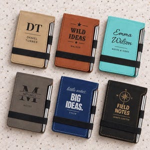 May include: Six rectangular pocket notebooks in tan, brown, teal, gray, blue, and black, each featuring a black elastic band, a silver pen, and unique custom text or graphics including names, compass designs, and phrases like Wild Ideas and Big Ideas.
