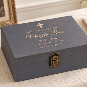 May include: A dark gray wooden keepsake box with a gold-toned metal latch. The lid features a gold-colored cross, the text "UNTIL WE MEET AGAIN", the name "Margaret Rose", the dates "1939 - 2024", and the phrase "Always in our prayers".