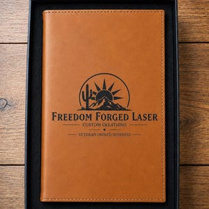 May include: A tan rectangular notebook with stitched edges, presented in a black box. The cover features a black laser-engraved logo with a cactus, sun, and mountains, reading Freedom Forged Laser, Custom Creations, and Veteran Owned Business.
