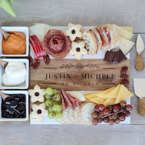 May include: A rectangular wooden serving board engraved with the text JUSTIN + MICHELE and 09.01.2019, surrounded by three square white bowls with wooden spoons, assorted cheeses, meats, crackers, grapes, and four metal serving utensils with wooden handles.