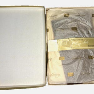 May include: An open, vintage box containing a pair of grey hosiery. The hosiery is wrapped in tissue paper and secured with a gold label that reads "Proportioned-Fit Ring olor Hosiery". The box is off-white.