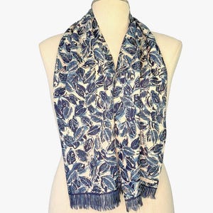 May include: A rectangular scarf featuring a repeating pattern of dark blue and light blue leaves on a cream-colored background. The scarf has a dark blue border along the bottom edges and is finished with a short, delicate fringe at both ends.