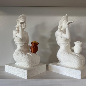 May include: Two white ceramic statues of women in traditional dress, each holding a small golden pot. The statues are seated with their hands raised to their faces, and they are placed on white pedestals with intricate designs.