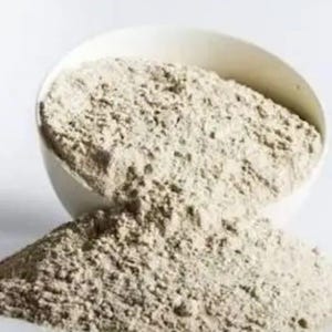 May include: A white ceramic bowl tipped over, spilling a pile of light brown powder. The powder has a fine, textured appearance, with a slightly uneven surface. The background is plain white, providing a clean contrast to the powder.