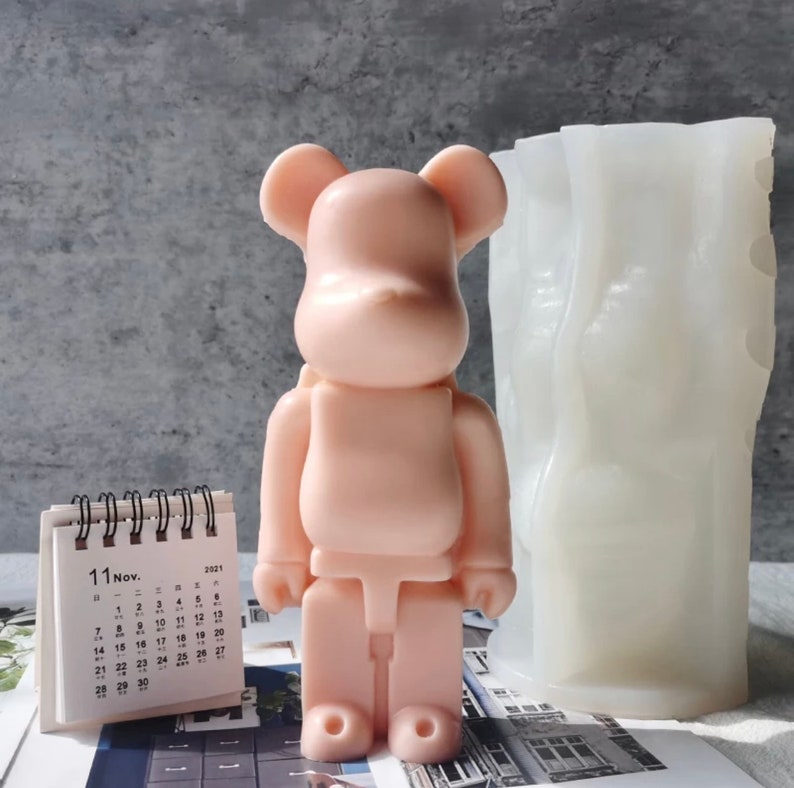 May include: A pink bear-shaped silicone mold and a white silicone mold for making candles. The bear mold has a simple design with no details. The other mold has a textured surface.