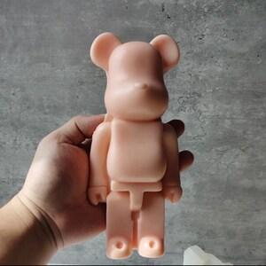 May include: A light pink, plastic bear-shaped toy with a simple design. The toy is standing upright and has a rounded head, small ears, and a small nose.