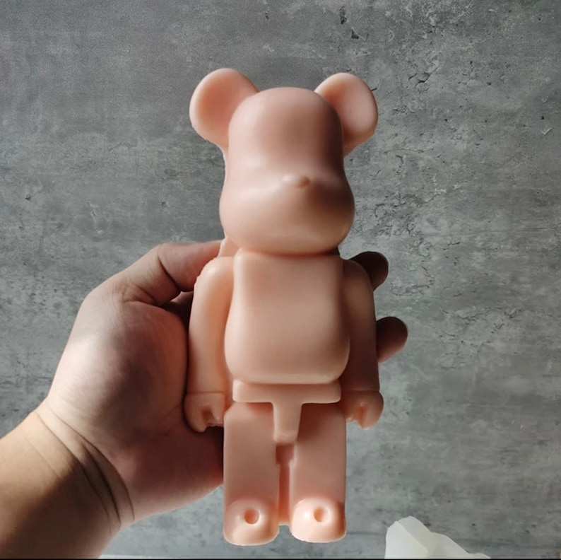 May include: A light pink, plastic bear-shaped toy with a simple design. The toy is standing upright and has a rounded head, small ears, and a small nose.