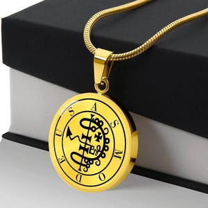 May include: Gold-toned pendant necklace with a circular charm. The charm features a black graphic design and the letters "S", "A", "S", "U", "E", "D", "O", and "M" around the edge. The necklace is displayed on a black and white box.