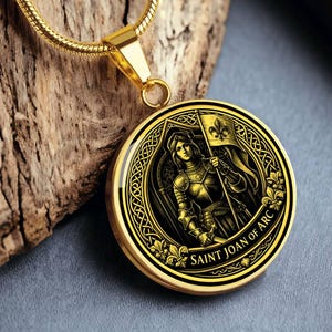 May include: Gold-toned pendant necklace featuring a detailed illustration of Saint Joan of Arc holding a flag with a fleur-de-lis. The pendant has a black and gold design with the text "SAINT JOAN OF ARC". The necklace is displayed on a wooden surface.