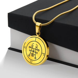 May include: Gold-colored pendant necklace with a circular charm. The charm features a black symbol within a yellow circle, with the letters "I", "S", "O", and "P" around the edge. The necklace is displayed on a black box.