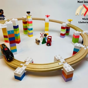 Könnte beinhalten: Ein Holz-Eisenbahnset mit bunten Bausteinen, einem Zug und Figuren. Die Schienen sind kreisförmig angeordnet und die Blöcke sind in verschiedenen Höhen gestapelt. Das Set beinhaltet einen rot-schwarzen Zug, eine braune Bärenfigur, eine Mädchenfigur und eine Jungenfigur. Der Text "Not included" und "Nicht im Lieferumfang" ist im Bild sichtbar.