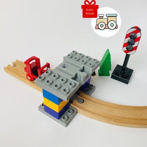 Op de afbeelding: Een speelgoedtreinset met een rode treinwagon, een grijze brug, een groene boom en een verkeerslicht. De brug wordt ondersteund door blauwe, gele en paarse blokken. De set bevat houten rails en een sticker met de tekst "Gratis Sticker".